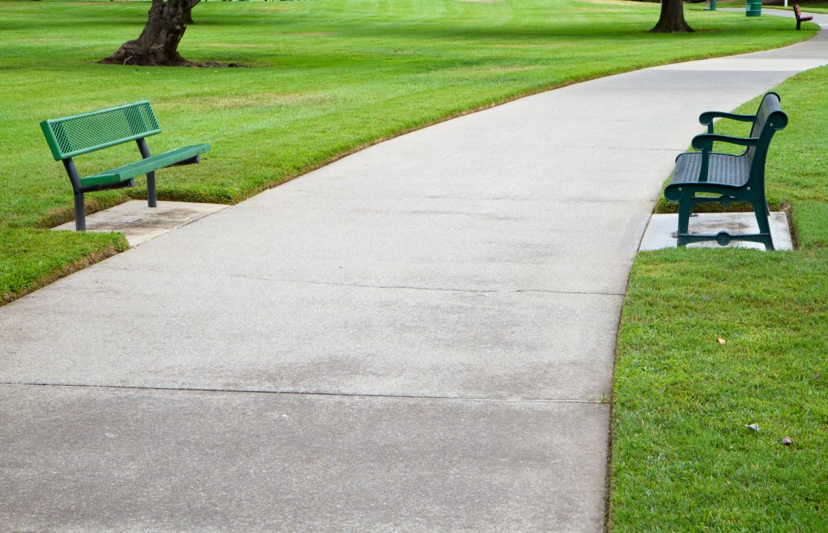 A winding concrete path with two green benches in a lush green park